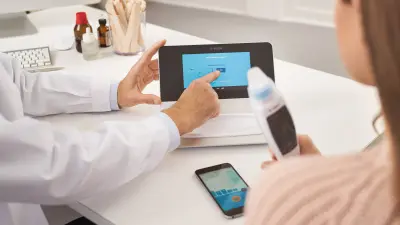 Patientin hebt Vivatmo-Messgerät in der Hand. Vivatmo pro auf Arztschreibtisch vor ihr liegend. Arzt deutet mit dem Finder aus den Screen.